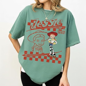 May include: A sage green short-sleeve t-shirt featuring a red graphic print of the character Jessie from Toy Story. The design includes the text Jessie and 1995, a red checkered pattern, and a line drawing of the character alongside a full-color image.