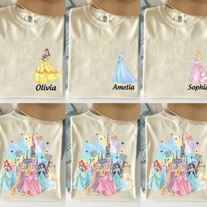 May include: Cream-colored t-shirts featuring princess designs. One shirt has a yellow dress with the name "Olivia", another has a blue dress with "Amelia", and a third has a pink dress with "Sophia". Other shirts feature a castle and multiple princesses.