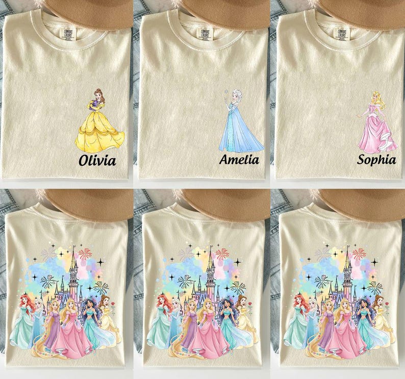 May include: Cream-colored t-shirts featuring princess designs. One shirt has a yellow dress with the name "Olivia", another has a blue dress with "Amelia", and a third has a pink dress with "Sophia". Other shirts feature a castle and multiple princesses.