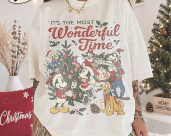 May include: An ivory short-sleeve t-shirt featuring a graphic print of Disney characters decorating a Christmas tree. The text reads, "IT'S THE MOST Wonderful Time." The design includes Mickey, Minnie, Donald, Daisy, Goofy, and Pluto in holiday attire.