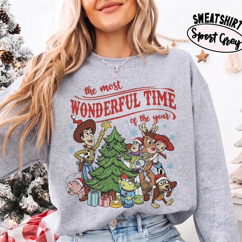 May include: A sport grey sweatshirt featuring a graphic print of Toy Story characters decorating a Christmas tree. The text reads, "the most WONDERFUL TIME of the year." The design includes Woody, Buzz Lightyear, Jessie, Rex, and other characters wearing Santa hats, surrounded by blue snowflakes and wrapped gifts.