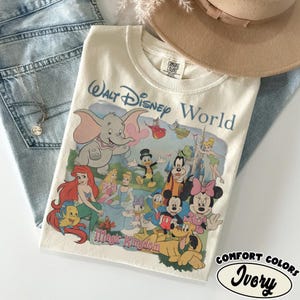 Puede incluir: Una camiseta de color marfil con un estampado gr&aacute;fico de Walt Disney World y Magic Kingdom que incluye varios personajes como Dumbo, Ariel, Flounder, Cenicienta, el Pato Donald, Goofy, Campanilla, Mickey Mouse, Minnie Mouse y Pluto.