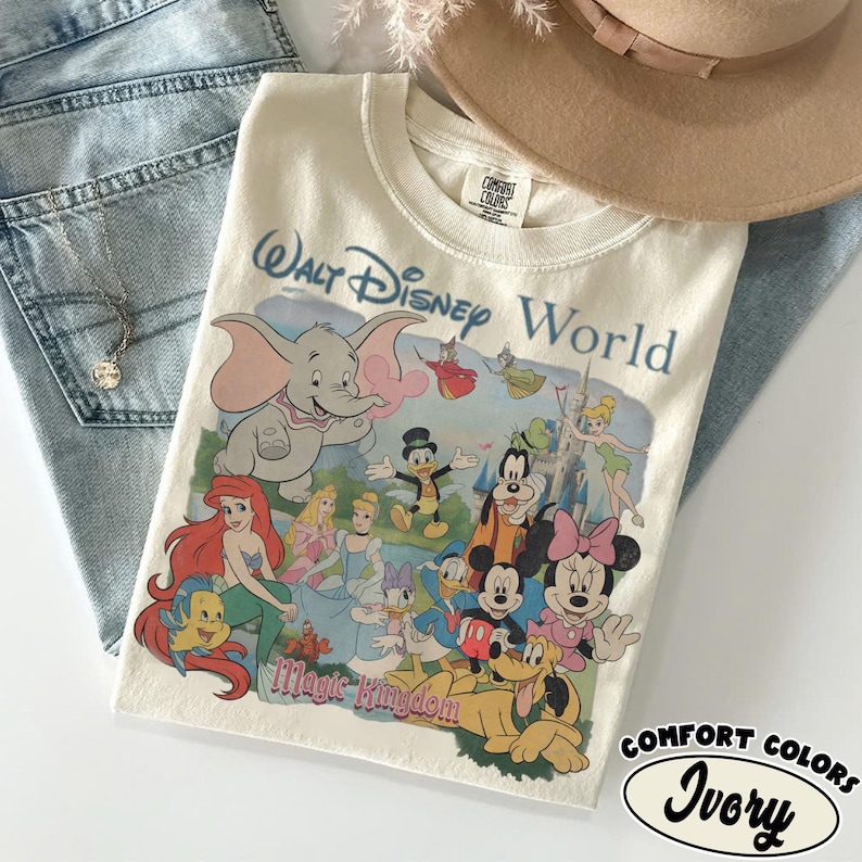 Puede incluir: Una camiseta de color marfil con un estampado gr&aacute;fico de Walt Disney World y Magic Kingdom que incluye varios personajes como Dumbo, Ariel, Flounder, Cenicienta, el Pato Donald, Goofy, Campanilla, Mickey Mouse, Minnie Mouse y Pluto.