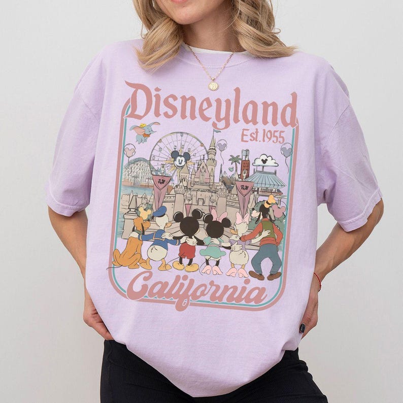 Puede incluir: Camiseta lila claro con un gr&aacute;fico de Disneyland. El dise&ntilde;o incluye las palabras "Disneyland California Est. 1955" junto con personajes de dibujos animados e im&aacute;genes del parque, como un castillo y una noria.