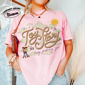Op de afbeelding: Een lichtroze T-shirt met korte mouwen voorzien van een afbeelding van een gele zon, beige wolken en de tekst "it's a Toy Story" in een touwachtig lettertype, met de zin "Baby, just say 'yes'" en kleine stripfiguren onder de tekst.