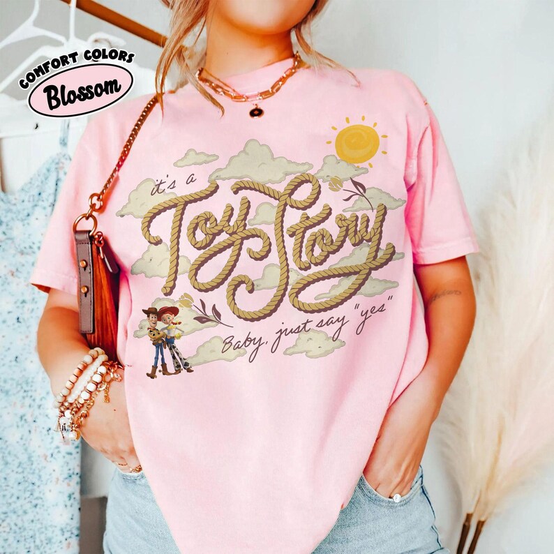 Op de afbeelding: Een lichtroze T-shirt met korte mouwen voorzien van een afbeelding van een gele zon, beige wolken en de tekst "it's a Toy Story" in een touwachtig lettertype, met de zin "Baby, just say 'yes'" en kleine stripfiguren onder de tekst.