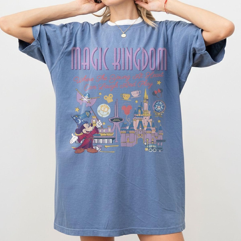 May include: A blue short-sleeve t-shirt featuring the text Magic Kingdom and a graphic print of a castle, a wizard character in a robe and hat, and various small icons including stars, a teacup, and a heart, all in shades of purple, yellow, and red.