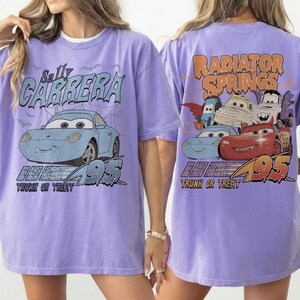 May include: A lilac t-shirt with a graphic of Sally Carrera from the film Cars, with the text "Sally Carrera" and "95". The back of the shirt shows "Radiator Springs" and other Cars characters in a Halloween theme.
