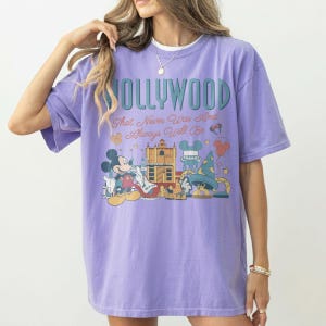 May include: A light purple t-shirt featuring a graphic print with the text HOLLYWOOD That Never Was And Always Will Be. The design includes a cartoon mouse character, a building, a wizard hat, and small star and balloon icons in muted colors.