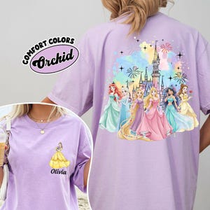 May include: A light purple t-shirt with a graphic of Disney princesses and a castle. The shirt has the words "Comfort Colors" and "Orchid" printed on it. The front of the shirt has a princess graphic and the name "Olivia".