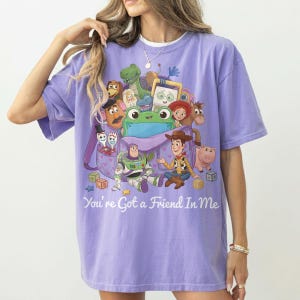May include: A light purple short-sleeve t-shirt featuring a graphic print of various animated characters from Toy Story emerging from a purple bag. The text "You're Got a Friend In Me" is printed in white below the characters and colorful toy blocks.