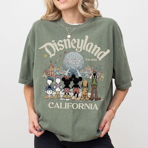 May include: An olive green t-shirt featuring the Disneyland logo with the text "Est. 1955" and "CALIFORNIA". The graphic includes cartoon characters like Mickey and Minnie Mouse, Donald and Daisy Duck, and Pluto in front of a futuristic sphere.