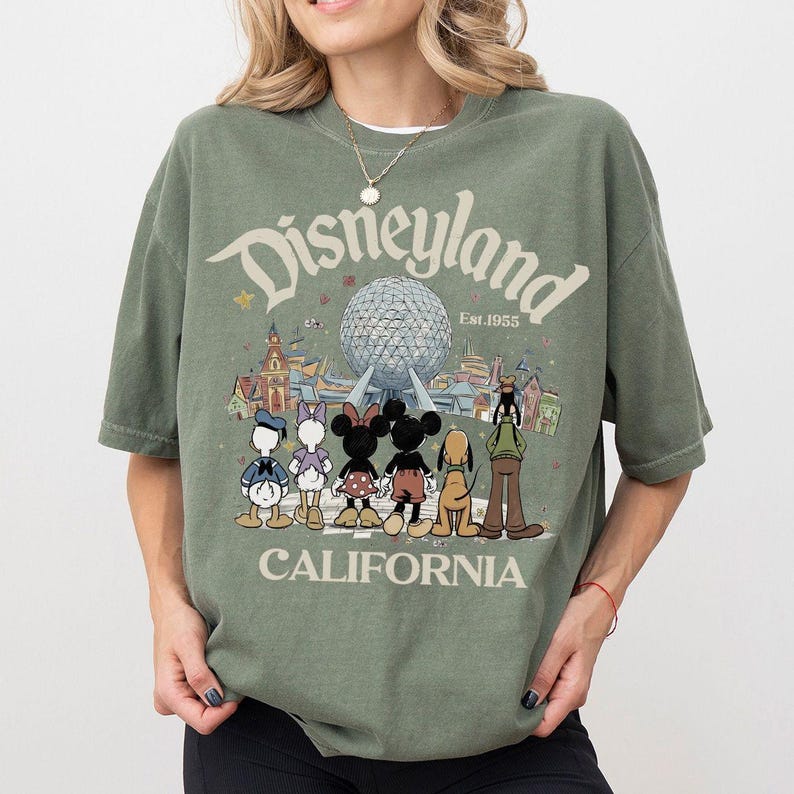 May include: An olive green t-shirt featuring the Disneyland logo with the text "Est. 1955" and "CALIFORNIA". The graphic includes cartoon characters like Mickey and Minnie Mouse, Donald and Daisy Duck, and Pluto in front of a futuristic sphere.