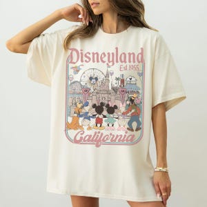 Puede incluir: Camiseta color crema con un gr&aacute;fico de Disneyland. El gr&aacute;fico incluye las palabras "Disneyland California Est. 1955" en escritura rosa, junto con personajes de dibujos animados e im&aacute;genes del parque. La camiseta tiene mangas cortas y un corte holgado.