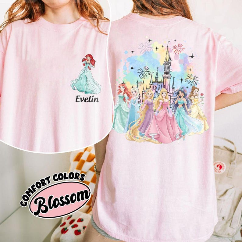 May include: A light pink t-shirt featuring a watercolor design of Disney princesses. The front of the shirt has a graphic of Ariel, with the name "Evelin" below. The back of the shirt features a castle and more princesses.