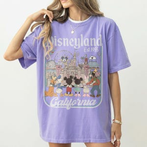 Puede incluir: Una camiseta de color lila con un gr&aacute;fico de Disneyland. El dise&ntilde;o incluye el logotipo de Disneyland, el texto "Est. 1955" e ilustraciones de Mickey Mouse, Minnie Mouse, Donald Duck y otros personajes de Disney. Tambi&eacute;n est&aacute; la palabra "California".