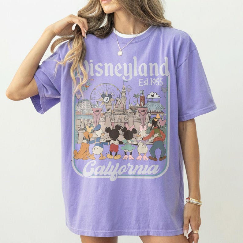 Puede incluir: Una camiseta de color lila con un gr&aacute;fico de Disneyland. El dise&ntilde;o incluye el logotipo de Disneyland, el texto "Est. 1955" e ilustraciones de Mickey Mouse, Minnie Mouse, Donald Duck y otros personajes de Disney. Tambi&eacute;n est&aacute; la palabra "California".