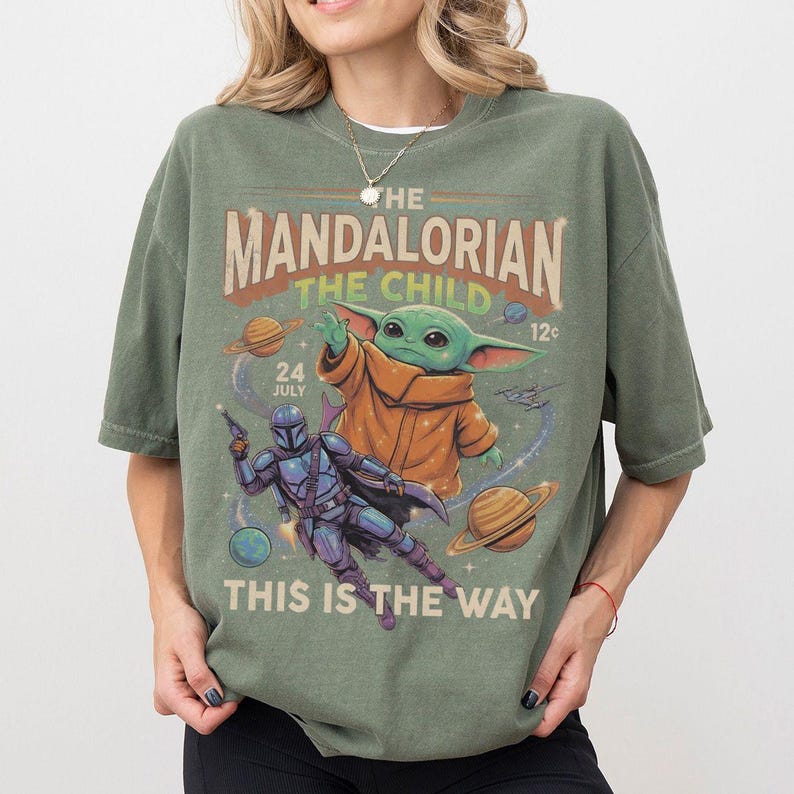 Op de afbeelding: Een olijfgroen t-shirt met een grafische print van The Mandalorian en The Child in de ruimte. Het ontwerp bevat planeten, sterren en de tekst "THE MANDALORIAN", "THE CHILD", "24 JULY", "12c" en "THIS IS THE WAY" in een vintage stijl.