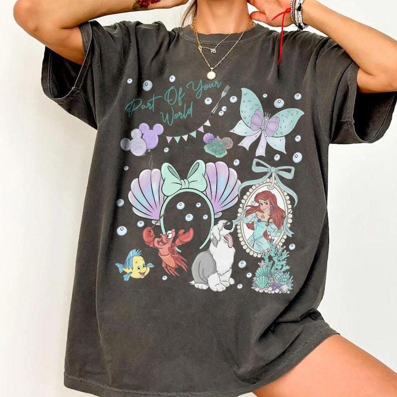 May include: A charcoal gray short-sleeve t-shirt featuring a graphic print with the text Part Of Your World. The design includes a mermaid, a crab, a fish, a dog, seashell ears, a butterfly, and bubbles in shades of teal, purple, and white.