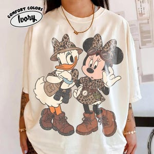 May include: An ivory t-shirt featuring a graphic of two cartoon characters wearing leopard print hats, vests, and brown hiking boots. One character wears a hat and carries binoculars, while the other wears goggles. Text in the corner reads COMFORT COLORS Ivory.