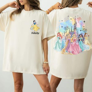 May include: Cream-colored t-shirt featuring a Snow White graphic with the word "Felicity" in black. The back of the shirt displays a colorful illustration of several princesses in front of a castle with fireworks.