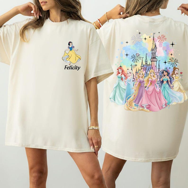May include: Cream-colored t-shirt featuring a Snow White graphic with the word "Felicity" in black. The back of the shirt displays a colorful illustration of several princesses in front of a castle with fireworks.