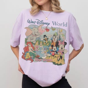 Puede incluir: Una camiseta de manga corta de color morado claro con un estampado gr&aacute;fico de varios personajes de Disney, incluidos Dumbo, Ariel, Cenicienta, Mickey Mouse, Minnie Mouse y el Pato Donald, con el texto Walt Disney World y Magic Kingdom.