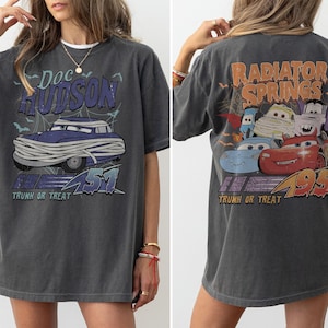 May include: Dark grey t-shirt with a graphic of Doc Hudson as a mummy, text reads "Doc Hudson 51 Trunk or Treat." The back shows "Radiator Springs 95 Trunk or Treat" with cartoon car characters in a Halloween theme.