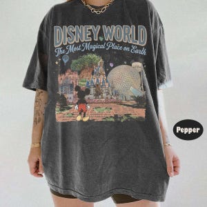 Puede incluir: Camiseta gris oscuro con un estampado colorido de Disney World con Mickey Mouse. El texto "DISNEY WORLD The Most Magical Place on Earth" est&aacute; impreso encima. La camiseta tiene mangas cortas y un corte holgado.