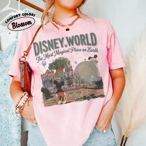 Puede incluir: Camiseta rosa claro con las palabras "DISNEY WORLD" y "The Most Magical Place on Earth" estampadas. El dise&ntilde;o incluye a Mickey Mouse, un castillo y una gran esfera. La camiseta tambi&eacute;n tiene las palabras "Comfort Colors" y "Blossom".