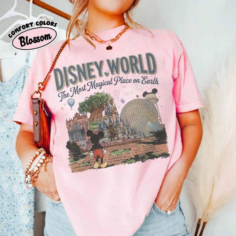 Puede incluir: Camiseta rosa claro con las palabras "DISNEY WORLD" y "The Most Magical Place on Earth" estampadas. El dise&ntilde;o incluye a Mickey Mouse, un castillo y una gran esfera. La camiseta tambi&eacute;n tiene las palabras "Comfort Colors" y "Blossom".