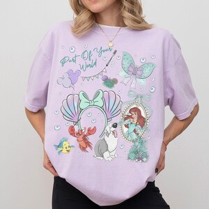 May include: A light purple short-sleeve t-shirt featuring a graphic print with the text Part Of Your World, a mermaid, a crab, a fish, a dog, a butterfly, and a pair of seashell ears. The design includes various bubbles and a banner.