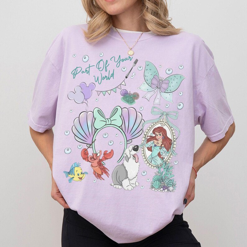 May include: A light purple short-sleeve t-shirt featuring a graphic print with the text Part Of Your World, a mermaid, a crab, a fish, a dog, a butterfly, and a pair of seashell ears. The design includes various bubbles and a banner.