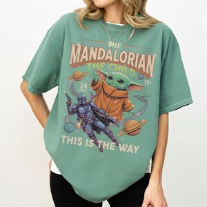 Op de afbeelding: Een saliegroen t-shirt met een grafische print van The Mandalorian en The Child in de ruimte. Het ontwerp bevat planeten, sterren en de tekst "THE MANDALORIAN THE CHILD", "24 JULY", "12" en "THIS IS THE WAY" in een vintage lettertype.