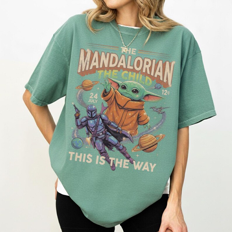 Op de afbeelding: Een saliegroen t-shirt met een grafische print van The Mandalorian en The Child in de ruimte. Het ontwerp bevat planeten, sterren en de tekst "THE MANDALORIAN THE CHILD", "24 JULY", "12" en "THIS IS THE WAY" in een vintage lettertype.