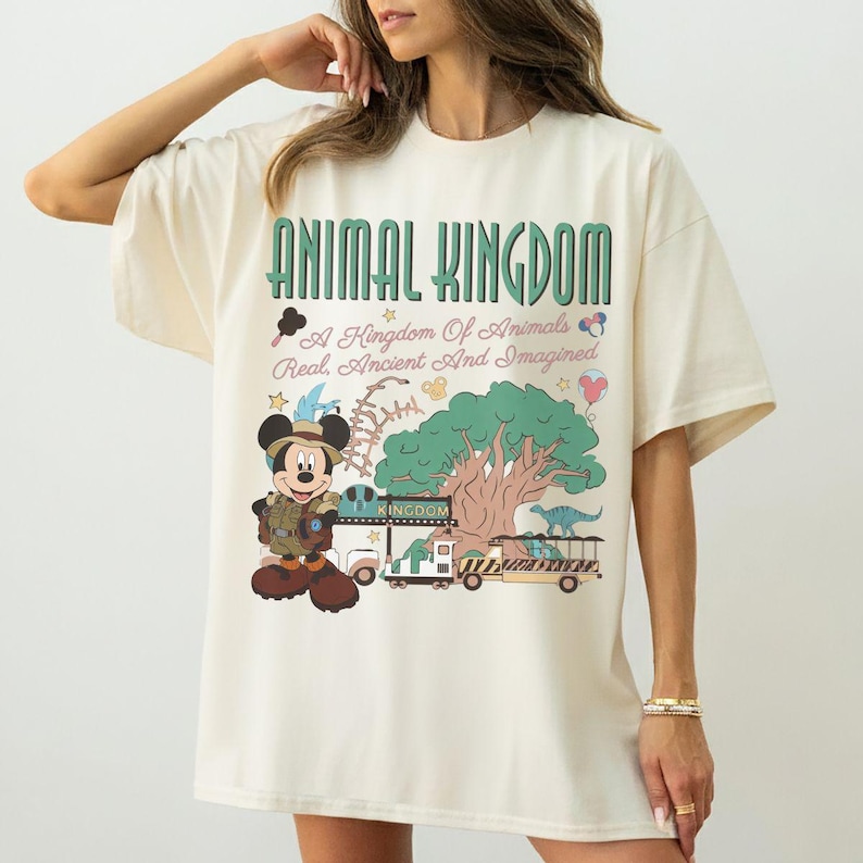 May include: An off-white short-sleeve t-shirt featuring a graphic print with the text ANIMAL KINGDOM, A Kingdom Of Animals Real, Ancient And Imagined. The design includes a cartoon mouse in safari gear, a large tree, and a safari vehicle.