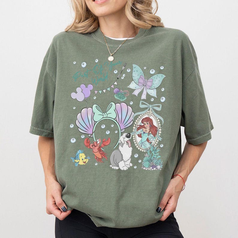 May include: An olive green t-shirt featuring a graphic print of a mermaid, a crab, a small yellow fish, a dog, a pair of seashell ears, a butterfly, and bubbles. The design includes a framed portrait of the mermaid and various decorative elements.