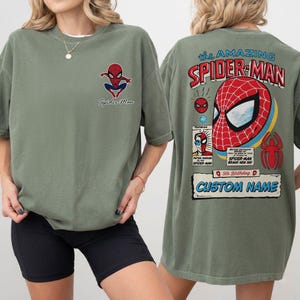 K&ouml;nnte beinhalten: Olivgr&uuml;nes T-Shirt mit Spider-Man-Grafiken auf der Vorder- und R&uuml;ckseite. Die Vorderseite zeigt ein kleines Spider-Man-Logo und Text. Die R&uuml;ckseite zeigt ein gro&szlig;es Spider-Man-Comic-Design mit dem Text "the AMAZING SPIDER-MAN".