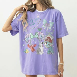May include: A lavender short-sleeve t-shirt featuring a graphic print with the text Part of Your World, a mermaid, a crab, a fish, a dog, a bow, and seashell motifs. The design includes various underwater elements and decorative accents.