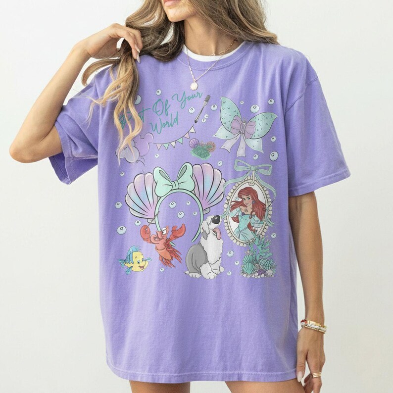 May include: A lavender short-sleeve t-shirt featuring a graphic print with the text Part of Your World, a mermaid, a crab, a fish, a dog, a bow, and seashell motifs. The design includes various underwater elements and decorative accents.