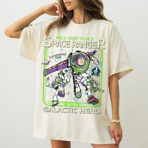 Op de afbeelding: Cr&egrave;mekleurig T-shirt met een afbeelding van Buzz Lightyear in een space ranger-uniform. Het ontwerp bevat de tekst "Buzz Lightyear's Space Ranger" en "Galactic Hero" in groen en wit. Opgericht in 1998.