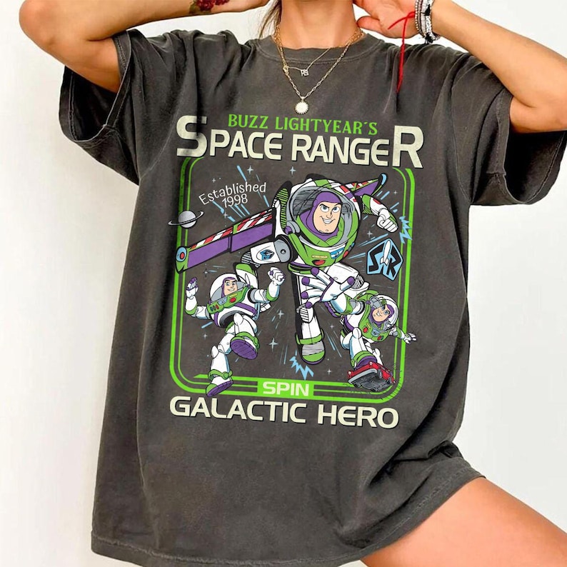 Op de afbeelding: Donkergrijs t-shirt met een afbeelding van Buzz Lightyear en de tekst "Buzz Lightyear's Space Ranger". Het ontwerp bevat groene en paarse accenten, met de woorden "Spin Galactic Hero" onderaan. Opgericht in 1998.