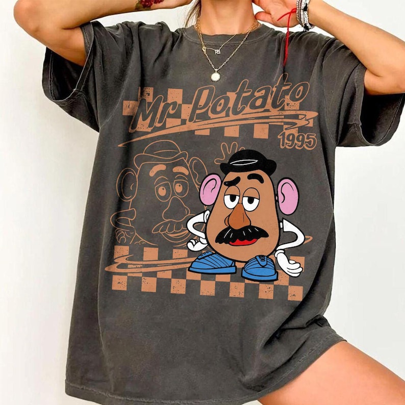May include: A charcoal gray short-sleeve t-shirt featuring a graphic of Mr. Potato Head with the text Mr. Potato 1995. The design includes a brown checkered pattern background, a line art outline of the character, and a full-color illustration.