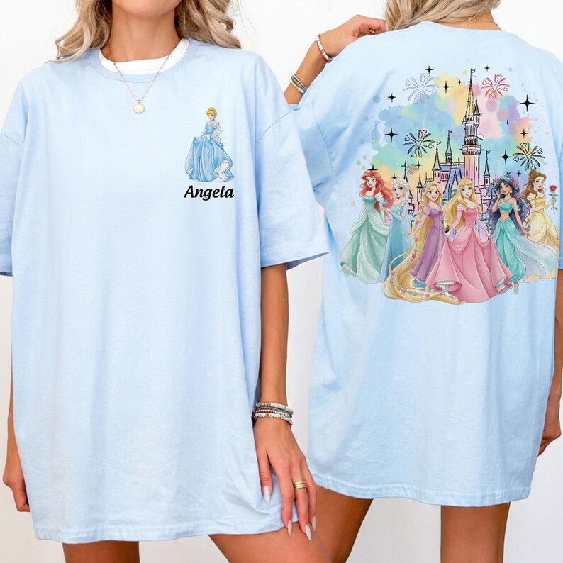 May include: Light blue t-shirts featuring a princess theme. The front of the shirt has a Cinderella graphic and the name "Angela". The back of the shirt has a castle and princess graphic. The shirt is a short-sleeved, crew neck style.