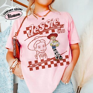 May include: A light pink short-sleeve t-shirt featuring a graphic print of the character Jessie from Toy Story. The design includes the text Jessie and 1995, a large portrait, a full-body illustration, and a brown and white checkered pattern.