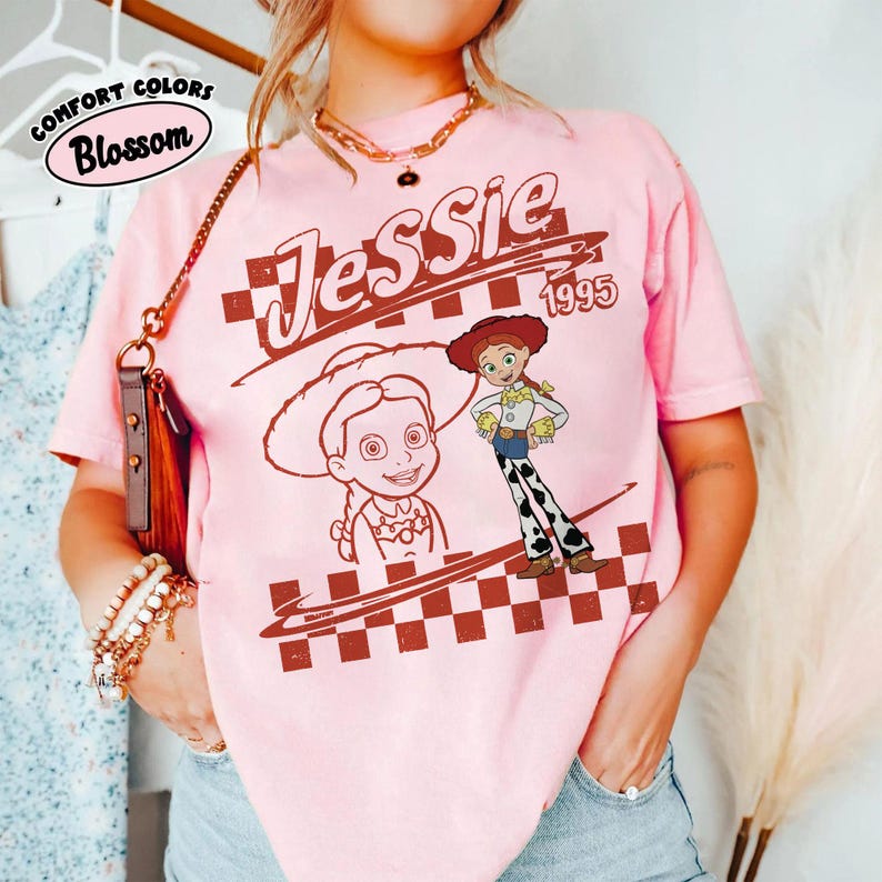 May include: A light pink short-sleeve t-shirt featuring a graphic print of the character Jessie from Toy Story. The design includes the text Jessie and 1995, a large portrait, a full-body illustration, and a brown and white checkered pattern.