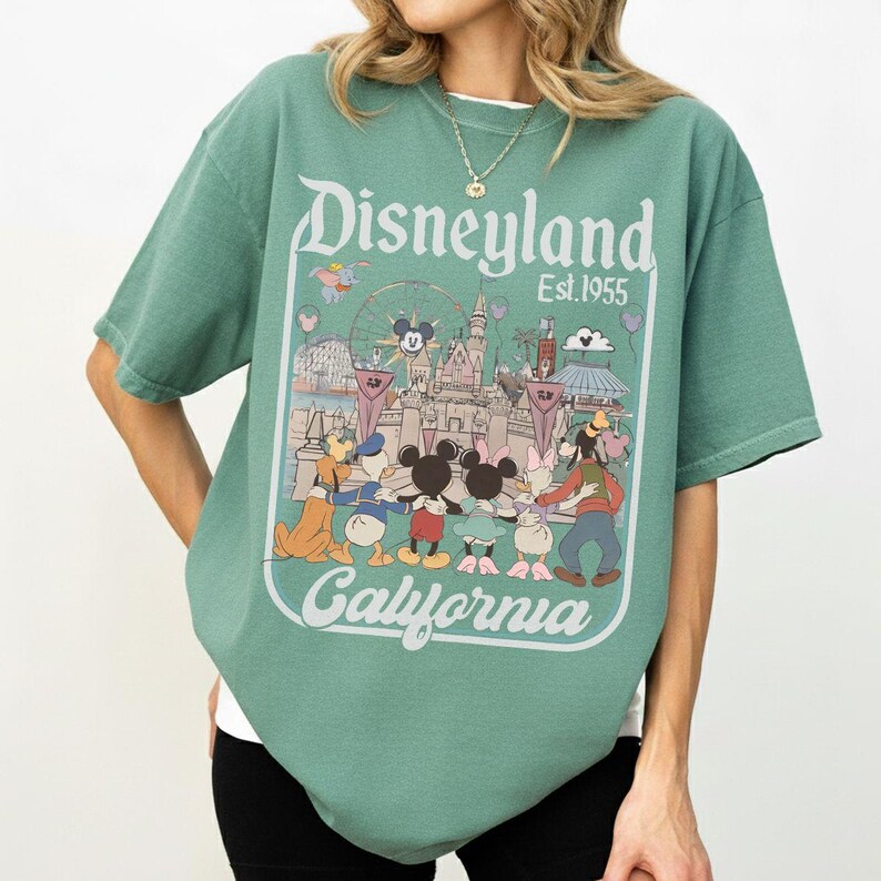 Puede incluir: Camiseta verde con un gr&aacute;fico de Disneyland. El gr&aacute;fico presenta el logotipo de Disneyland, el texto "Est. 1955" y una colorida ilustraci&oacute;n de personajes de Disney y atracciones del parque. La camiseta es de corte holgado.