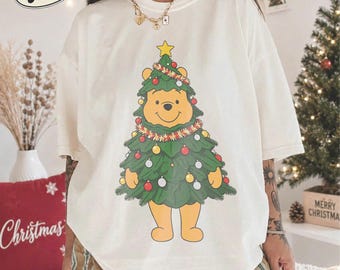 May include: An ivory short-sleeve t-shirt featuring a graphic of a yellow bear dressed as a Christmas tree. The bear is adorned with green pine branches, a yellow star on top, and colorful ornaments. The shirt has a crew neckline and a relaxed fit.