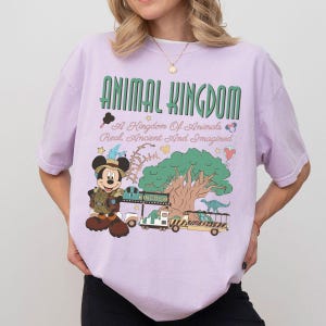 May include: A light purple t-shirt featuring a graphic print with the text ANIMAL KINGDOM A Kingdom Of Animals Real, Ancient And Imagined. The design includes a cartoon mouse in safari gear, a large tree, a safari vehicle, and a dinosaur.