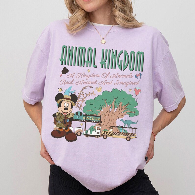May include: A light purple t-shirt featuring a graphic print with the text ANIMAL KINGDOM A Kingdom Of Animals Real, Ancient And Imagined. The design includes a cartoon mouse in safari gear, a large tree, a safari vehicle, and a dinosaur.